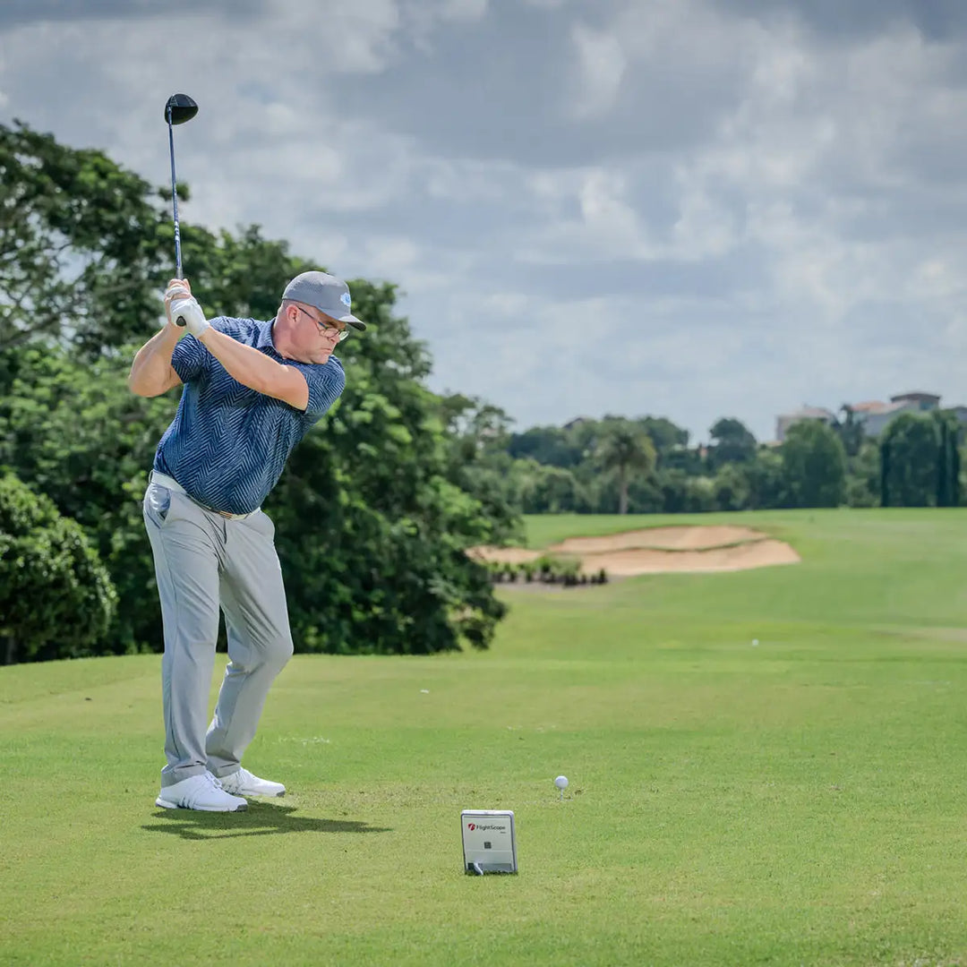Male golfer swinging a golf club on a lush green fairway with a golf launch monitor positioned nearby.